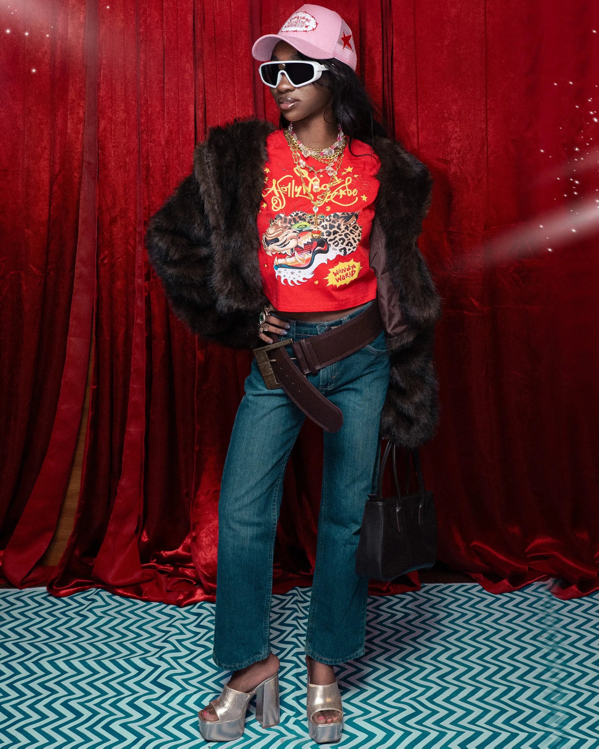 Person wearing a red graphic sweatshirt, blue jeans, and a fur coat against a red curtain background.